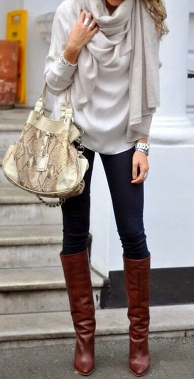 Woman on the sidewalk wearing a pale silver sweater, beige scarf, blue jeans and brown boots with a snakeskin handbag