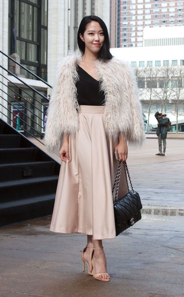 Asian woman wearing a black top with a nude midi skirt, nude heels, and a fluffy coat