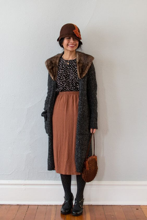 Woman wearing a polka dotted top, rust colored midi skirt, long coat, and a 20s-style hat