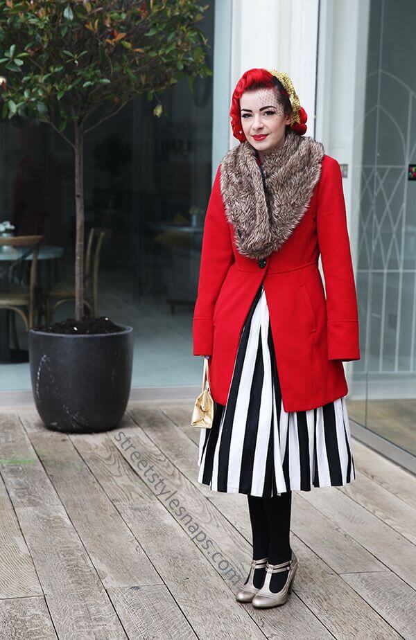 Woman wearing a striped skirt, red coat, fur collar, and a fascinator