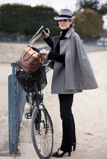 Woman with a bicycle wearing black pants, a grey cape coat, and a grey fedora