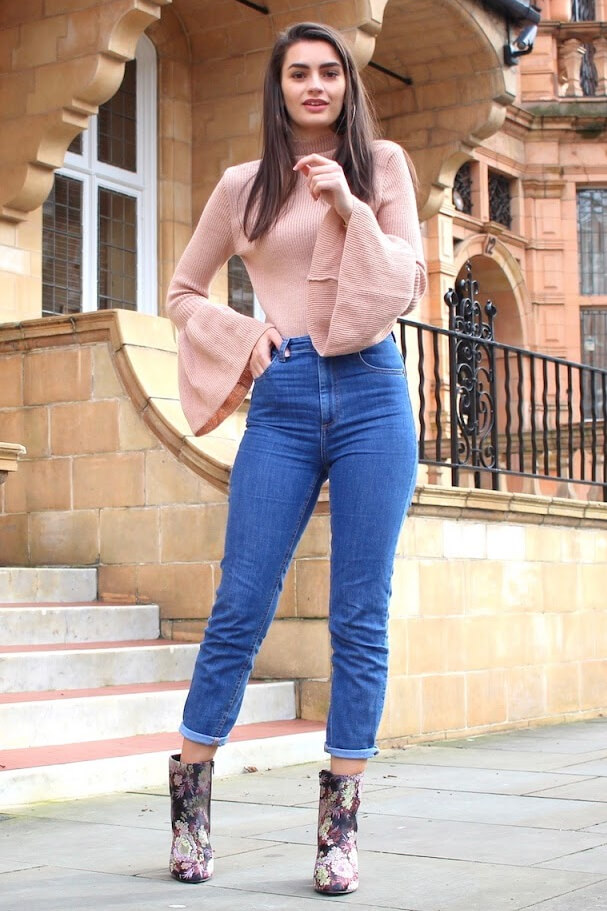 Fashionable brunette is wearing high-waisted jeans and a blush pink woolen bell sleeve sweater. Ease yourself into spring in a turtleneck bell sleeve sweater and trendy high-waisted mom jeans.