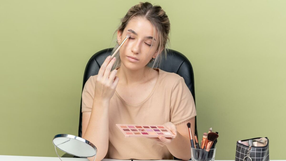 with closed eyes young beautiful girl sitting at desk with makeup tools applying eyeshadow with makeup brush isolated on olive green background