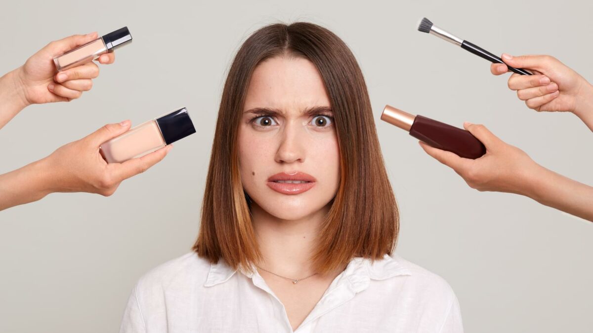 Young attractive girl with dark hair doing make-up being surrounded with hands with brushes for make-up, expressing shock and negative emotions.