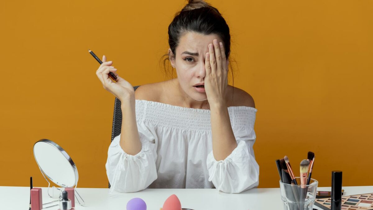 sad beautiful girl sits at table with makeup tools closes eye with hand holding eyeliner looking at camera isolated on orange background with copy space