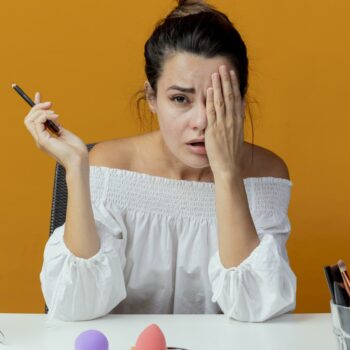 sad beautiful girl sits at table with makeup tools closes eye with hand holding eyeliner looking at camera isolated on orange background with copy space