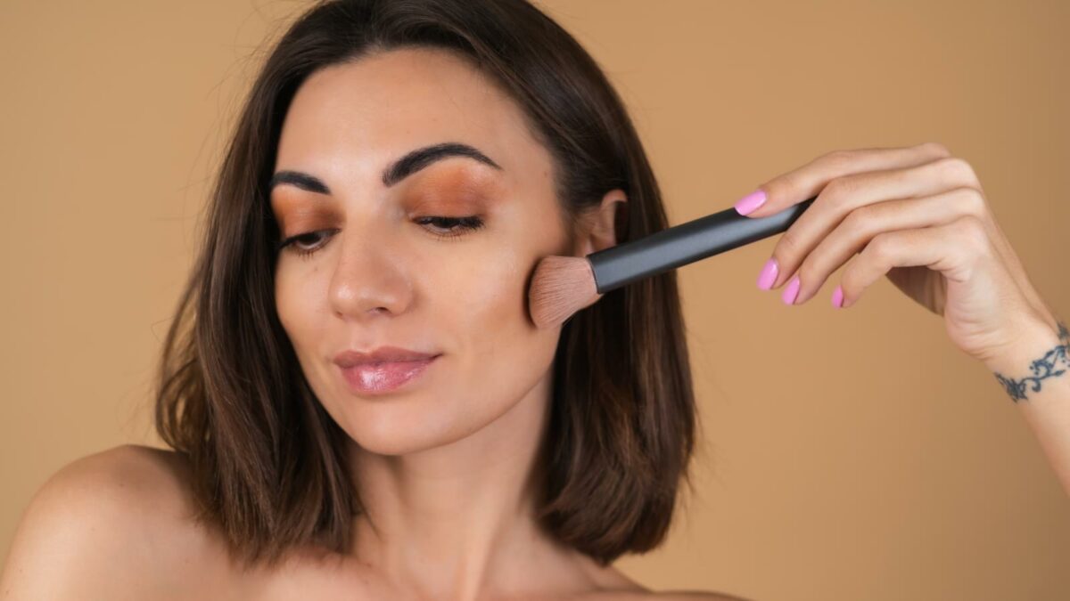 Portrait of a young woman on a beige background with natural warm make-up and smooth clean skin, holding a brush for blush
