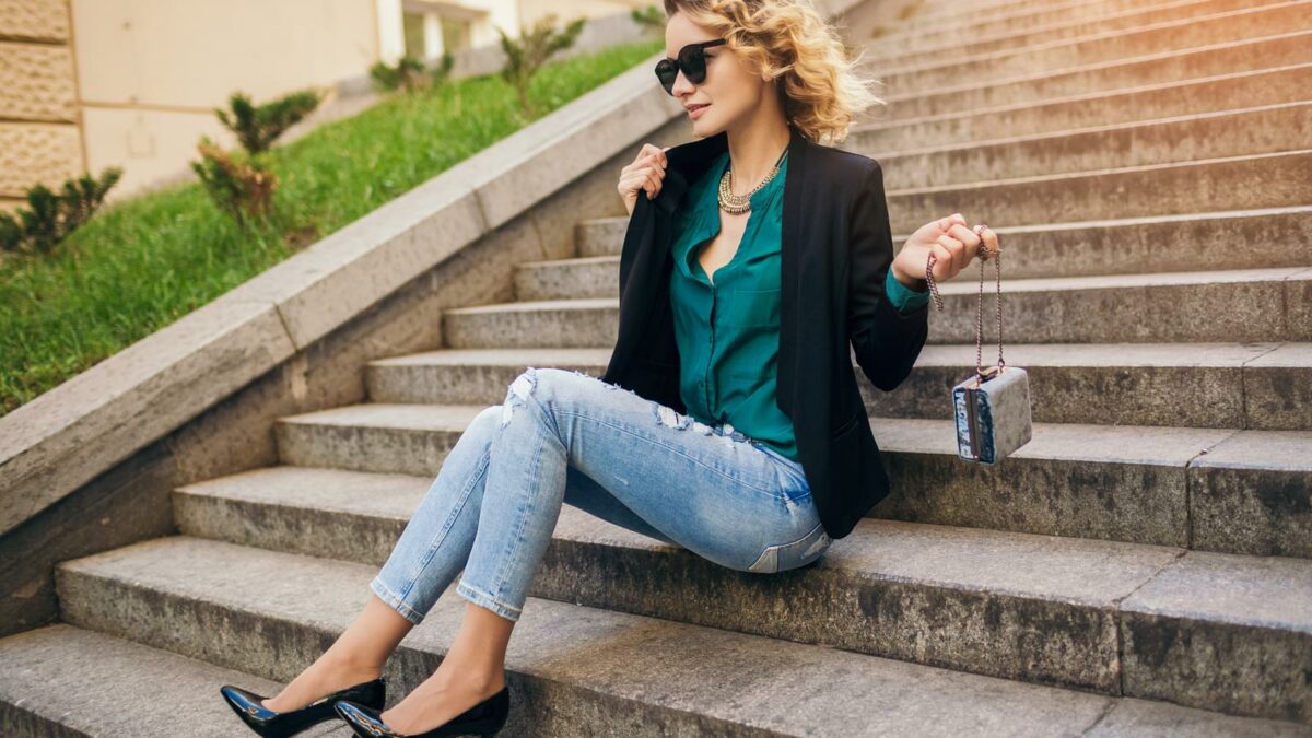 young stylish beautiful woman sitting in street, wearing jeans, black jacket, green blouse, sunglasses, holding purse, elegant style, summer fashion trend, legs details, high heeled shoes, footwear