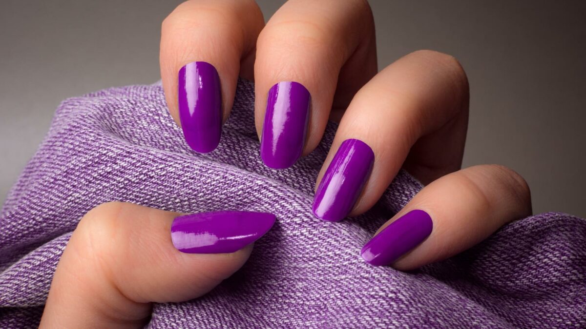 The female hand with purple nails is holding purple denim textile on gray background. Manicure concept.