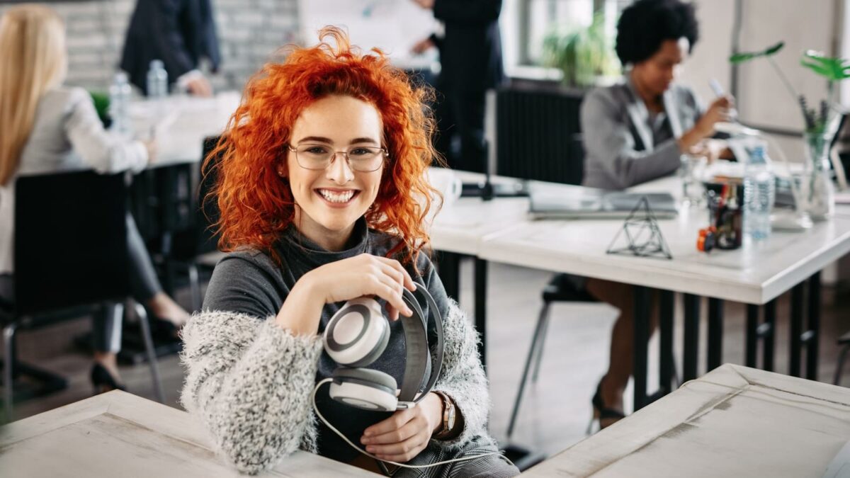 Young happy redhead businesswoman holding headphones and looking at camera while sitting at her desk in the office. Her colleagues are working in the background.