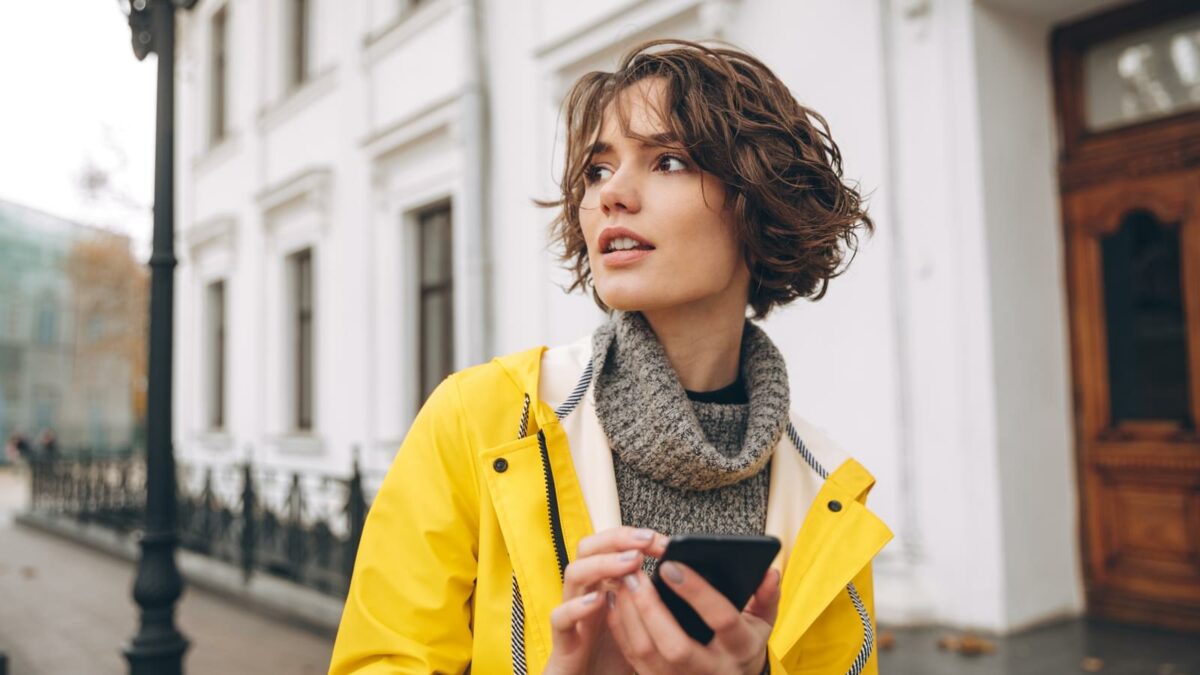 Image of beautiful young woman dressed in raincoat walking outdoors chatting by mobile phone. Looking aside.