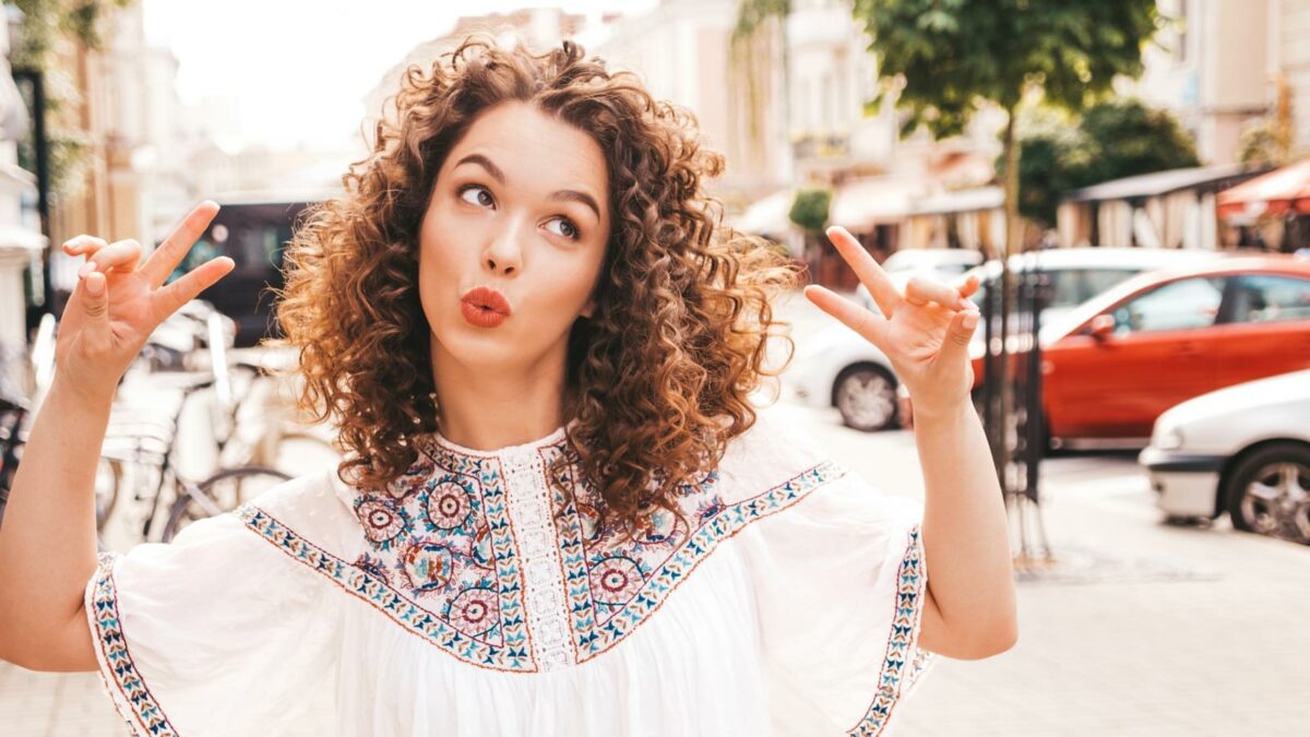 Portrait of beautiful smiling model with afro curls hairstyle dressed in summer hipster white dress.Sexy carefree girl posing in the street background.Trendy funny and positive woman shows peace sign