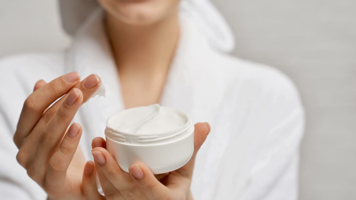Close up hands of young woman holding white box with moisturizer and showing at camera. Positive model in white bathrobe with pleasure demonstrates cream. Concept of skin care and beauty.