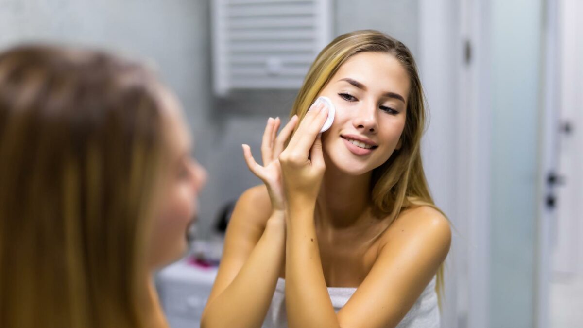 smiling young woman applying lotion to cotton disc for washing her face at bathroom