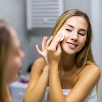 smiling young woman applying lotion to cotton disc for washing her face at bathroom