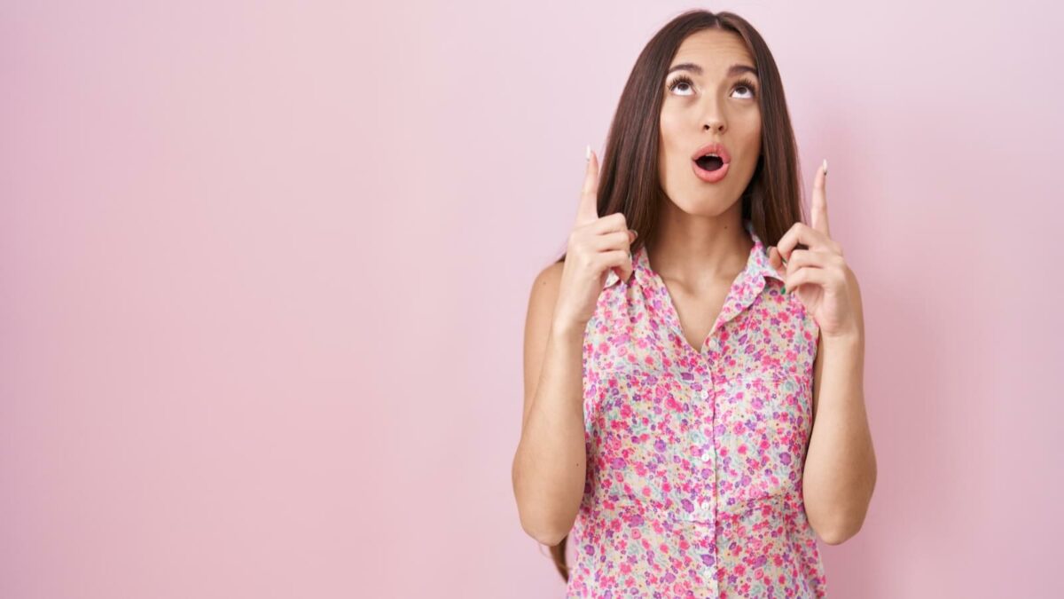 Young hispanic woman with long hair standing over pink background amazed and surprised looking up and pointing with fingers and raised arms.