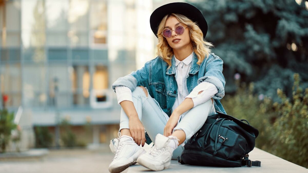 Modern lifestyle woman sitting outdoors with glass building in background. Cool young modern Caucasian female model wearing fashionable jeans, clothes, round glasses, hat and backpack.