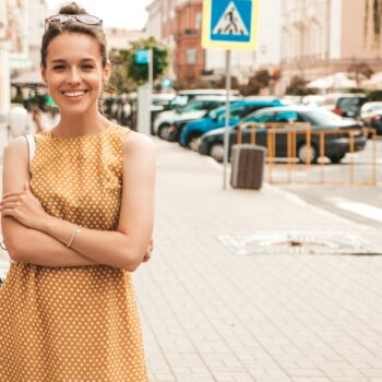 Portrait of beautiful smiling hipster model dressed in summer yellow dress. Trendy girl posing in the street background. Funny and positive woman having fun