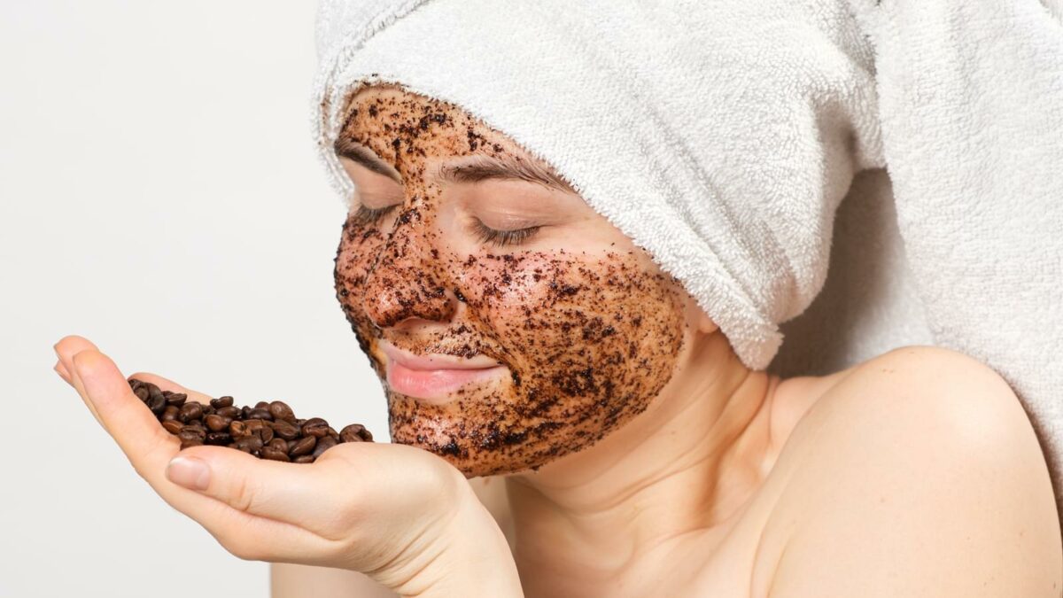 A woman with a coffee mask or scrub on her face holds a handful of coffee beans in her hand.