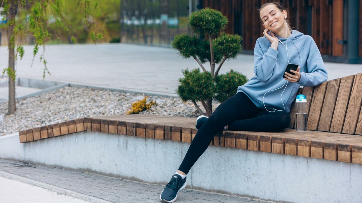 Side view of girl sitting on leg on bench, listening to music. Pretty young female smiling, with closed eyes, enjoying, wearing casual clothes. Concept of urban lifestyle,