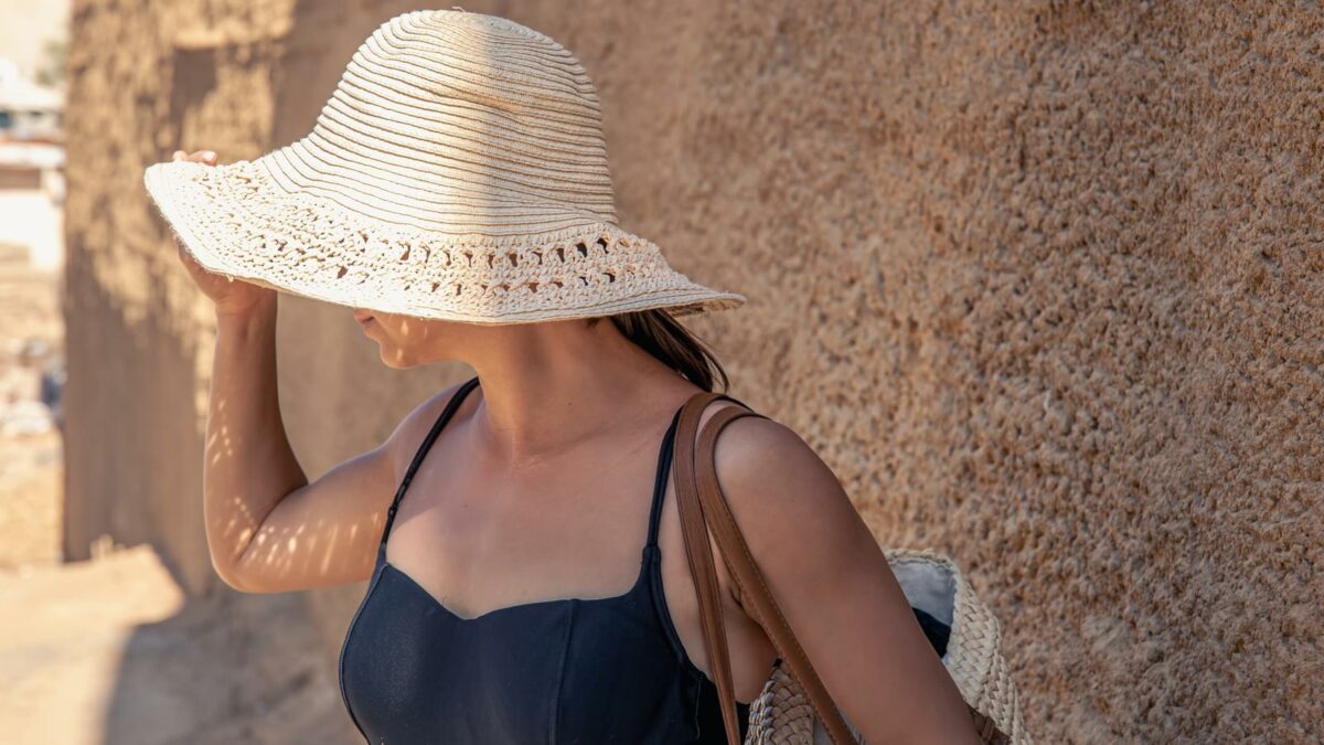 A young woman in a large straw hat hides from the sun near a sandy wall on a hot day.