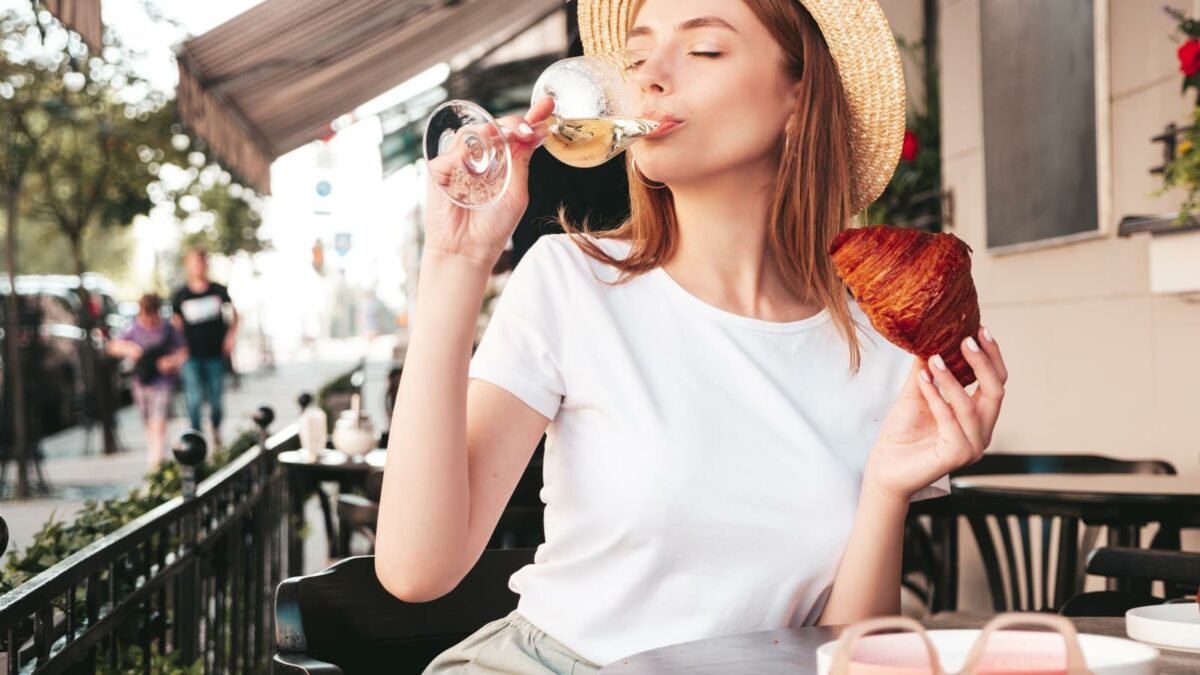 Young beautiful smiling hipster female in trendy summer clothes.Carefree woman posing at veranda cafe in the street.Positive model drinking white wine.Enjoying vacation.Eating croissant. In hat