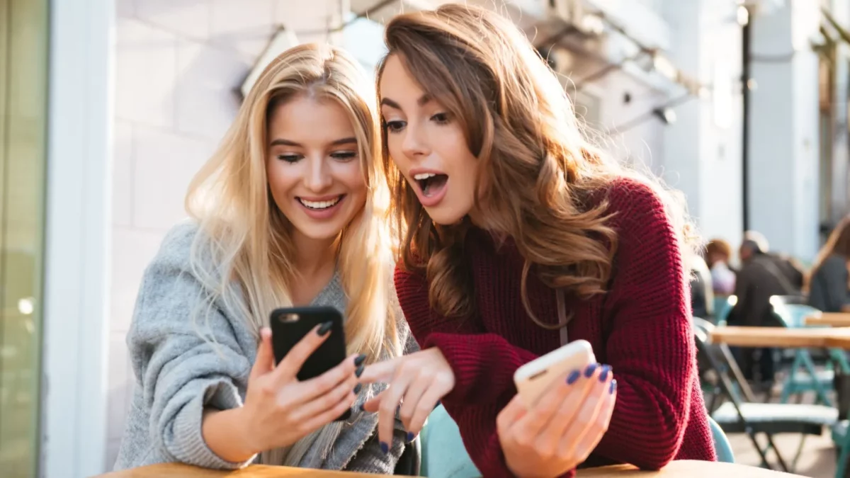 Two excited young girls using mobile phones while sitting at the cafe outdoors